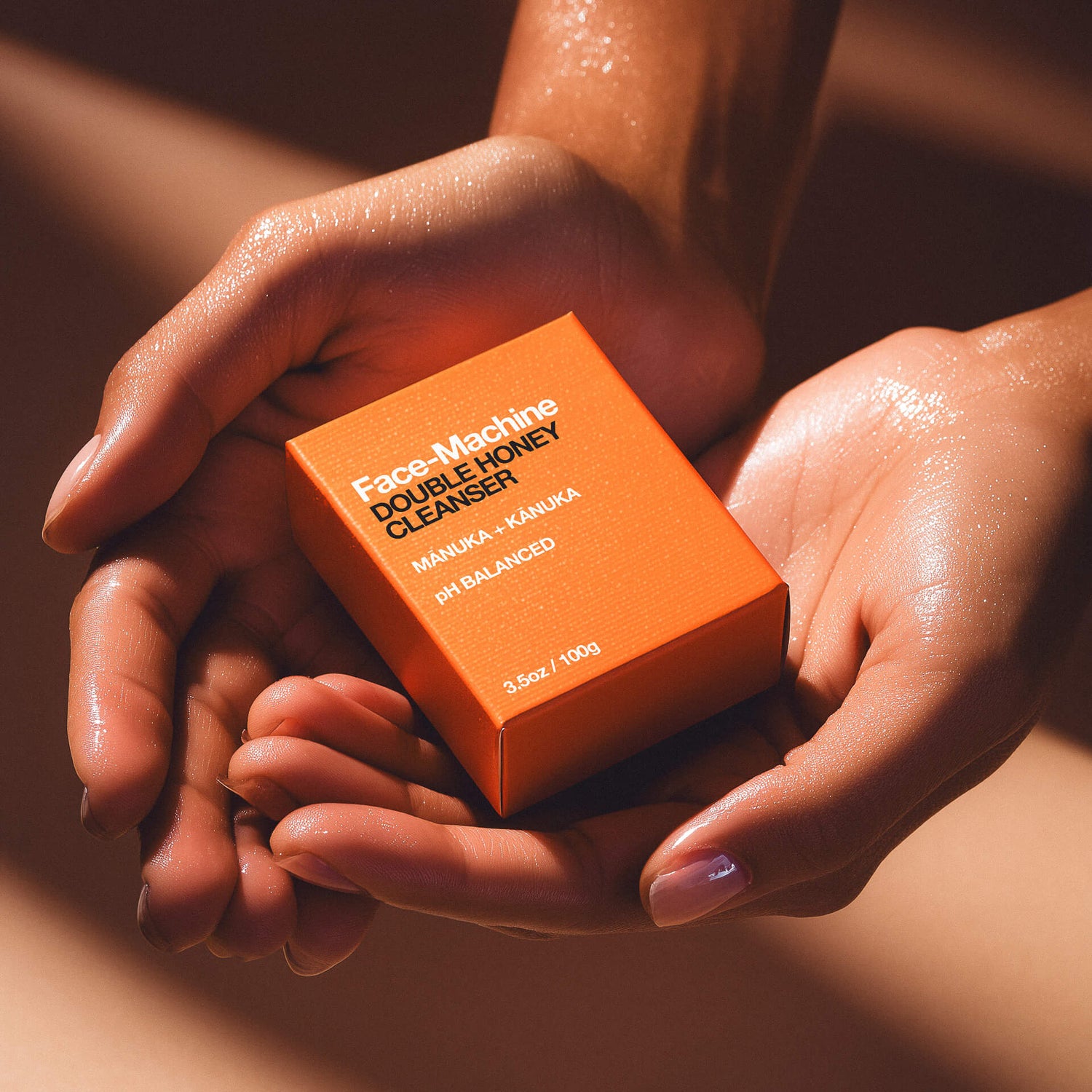 Orange box of 'Face Machine Double Honey Cleanser' held between hands with a warm background. facial cleanser, clean skin, skin repair, gift for wife, skincare routine, gentle face wash, hydrating cleanser, anti-aging skincare, daily cleanser, sensitive skin care, skin revitalization, luxury skincare gift, natural cleanser, deep pore cleanse, skin renewal, manuka honey